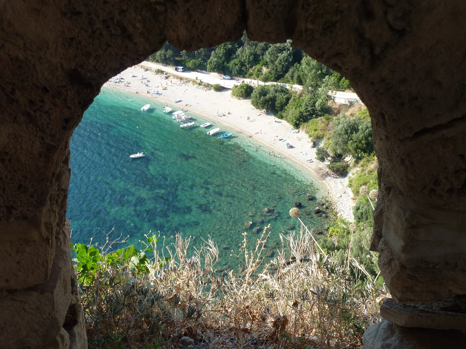 View of a long, curved beach with blue water seen through an arched stone window of a castle.