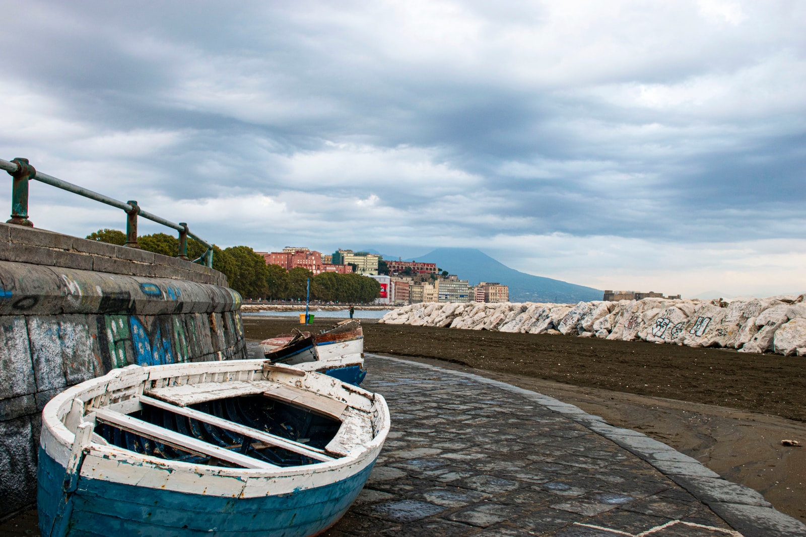An old wooden rowboat rests on the stone walkway of Via Caracciolo, with a misty, romantic view of Mount Vesuvius in the background.