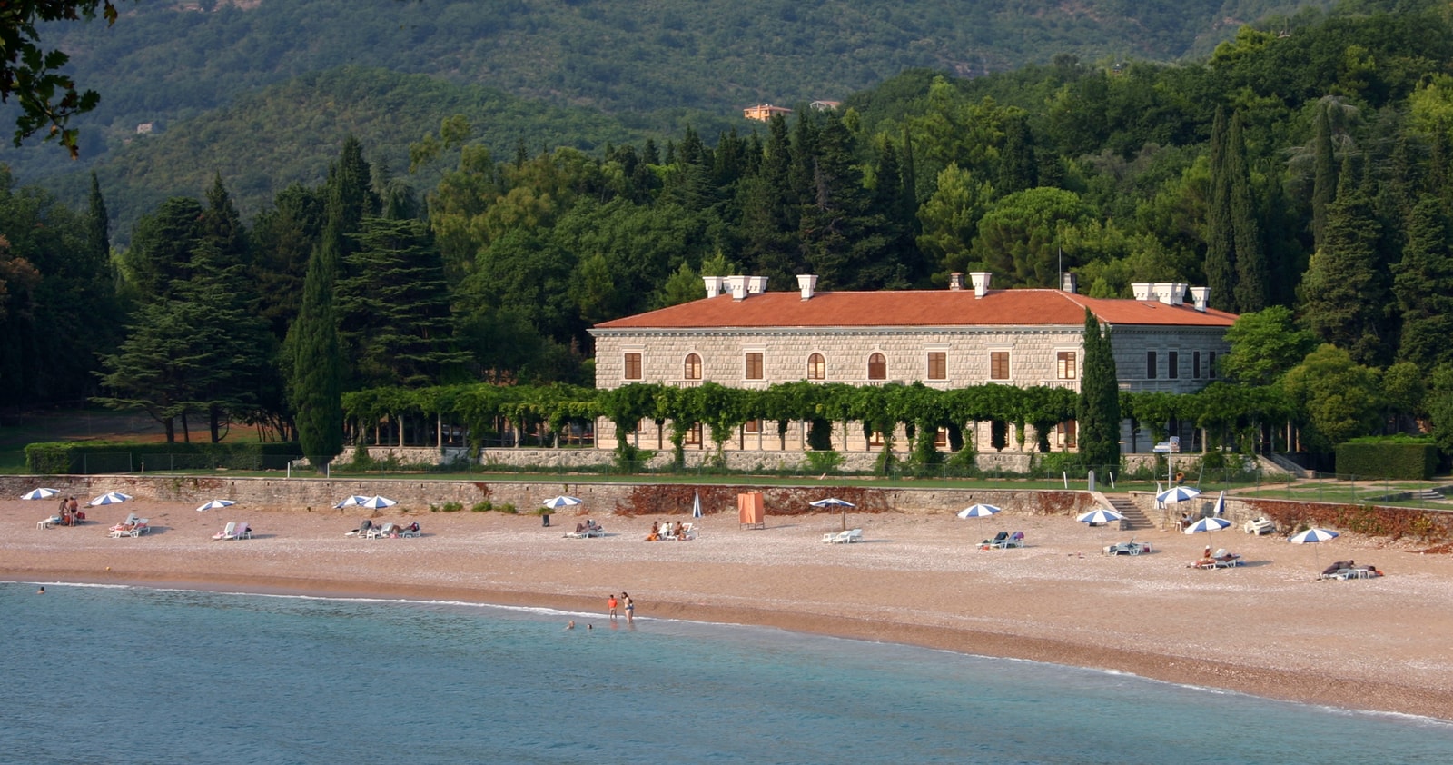 A stone villa with a terracotta roof sits beside a serene beach cove with calm, clear water.