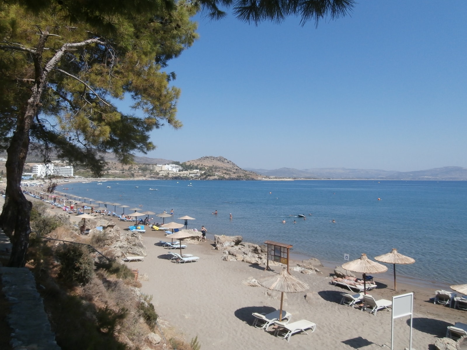 A calm view of Vlycha Beach shows a sand-and-pebble shore with sun loungers and trees framing the bay.