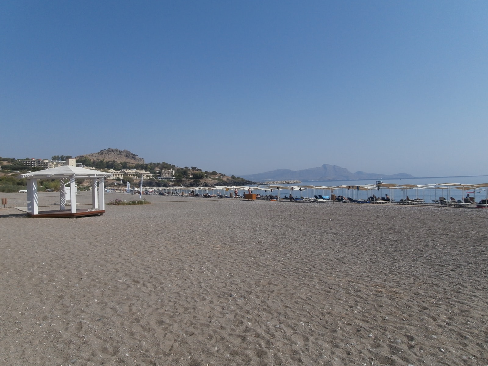 A wide view of Vlycha Beach shows its open shoreline with a quay and distant hills.