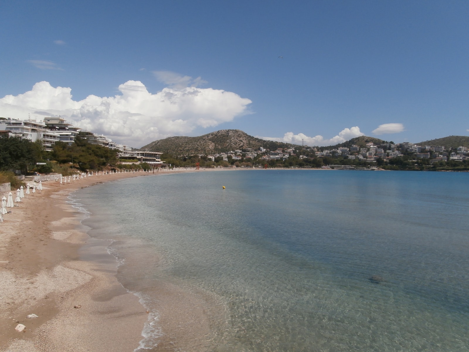 A wide view of Vouliagmeni's curved bay, filled with turquoise water and lined with beach umbrellas.