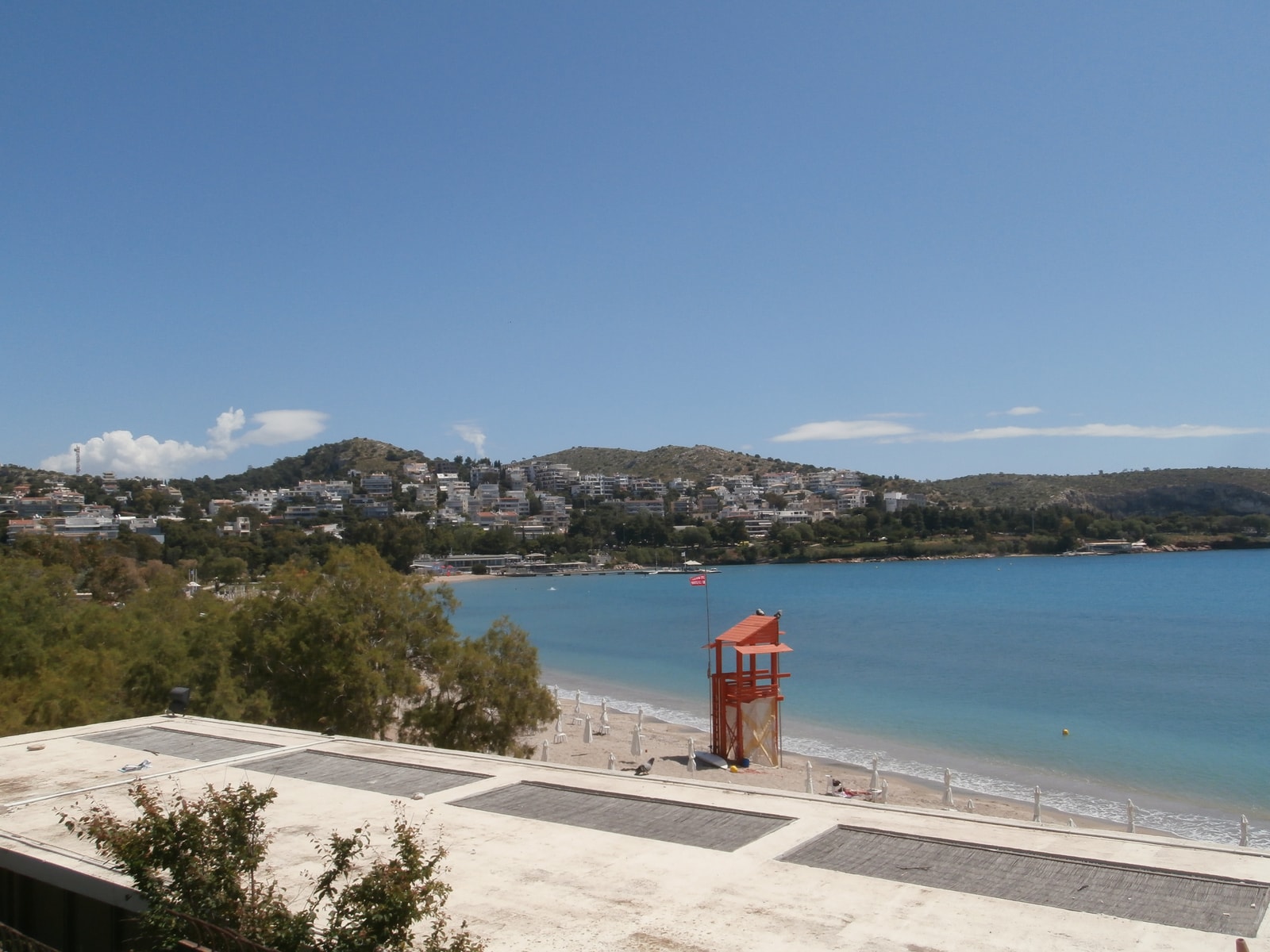 The shoreline of Vouliagmeni with a lifeguard tower, beach facilities, and green hills in the background.