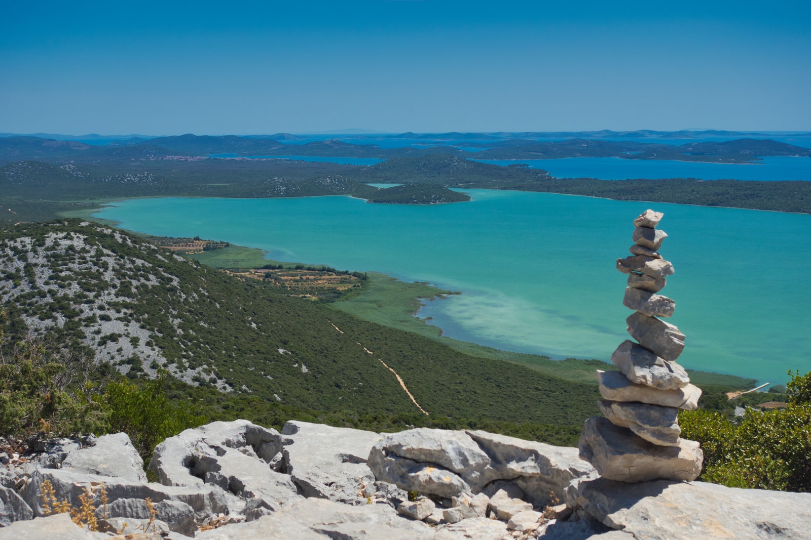 A panoramic view of the vast, turquoise Vrana Lake from the Kamenjak viewpoint, with a stone cairn in the foreground.