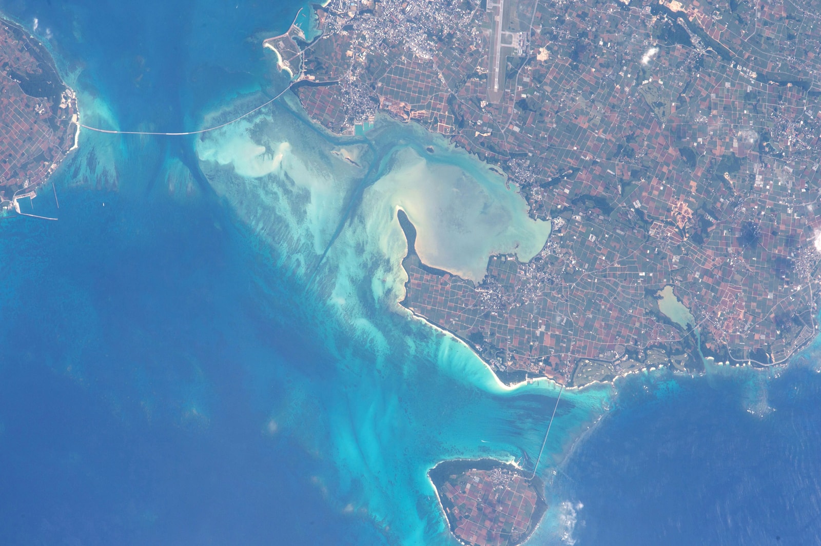 An aerial view of a bay with light blue and green water, showing sandbars and a bridge.