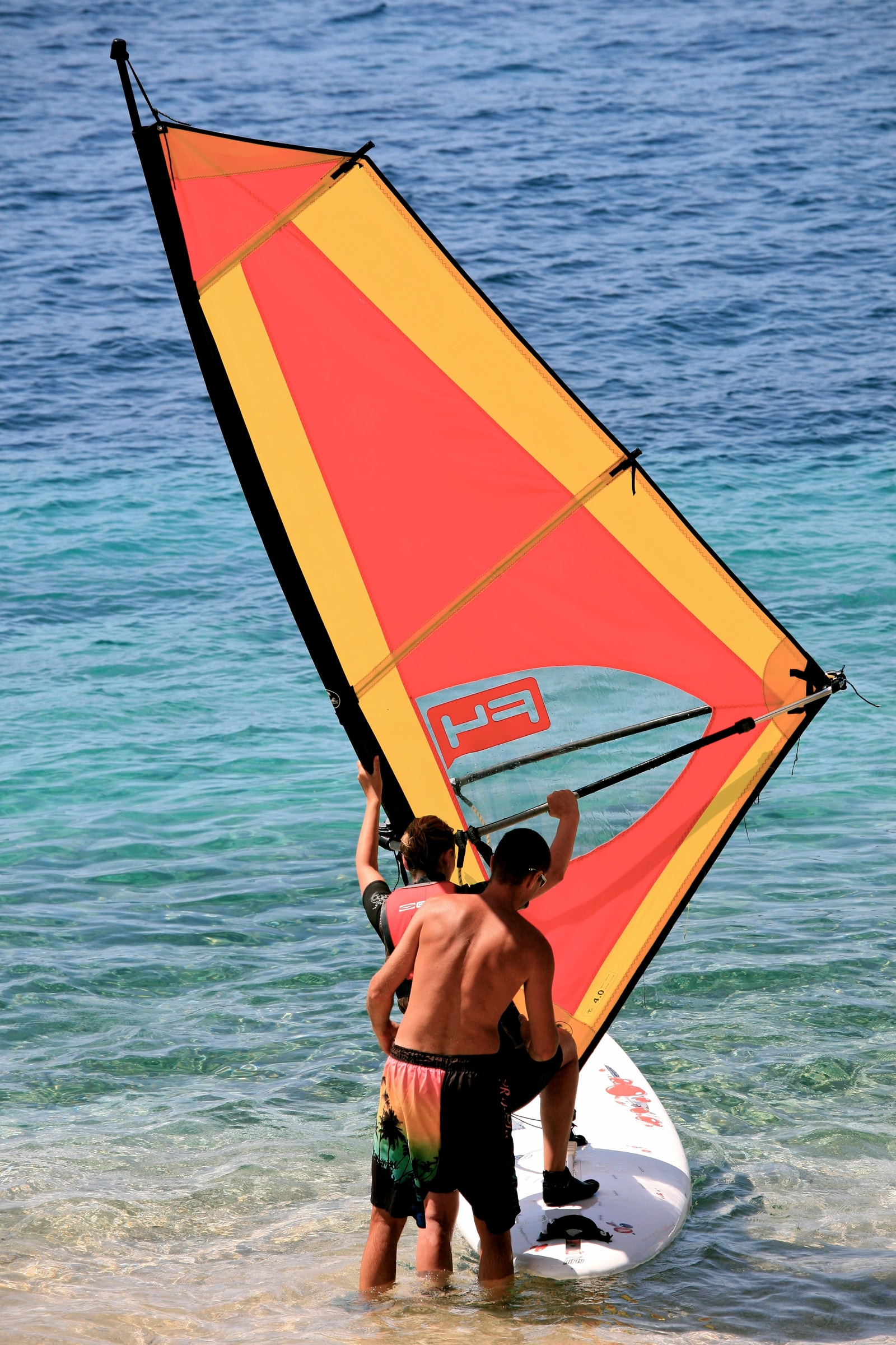 A windsurfing instructor teaches a beginner in the shallow, clear water near the shore.