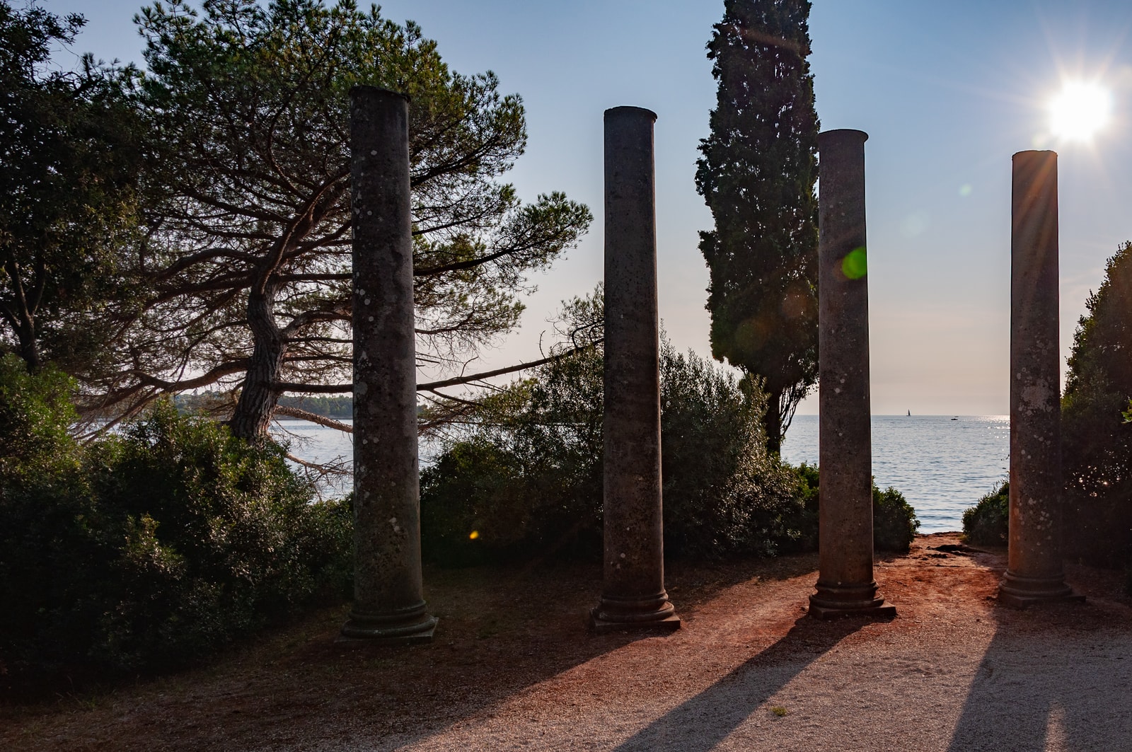 Four ancient stone columns stand among pine and cypress trees, overlooking the sea as sunlight filters through the branches.