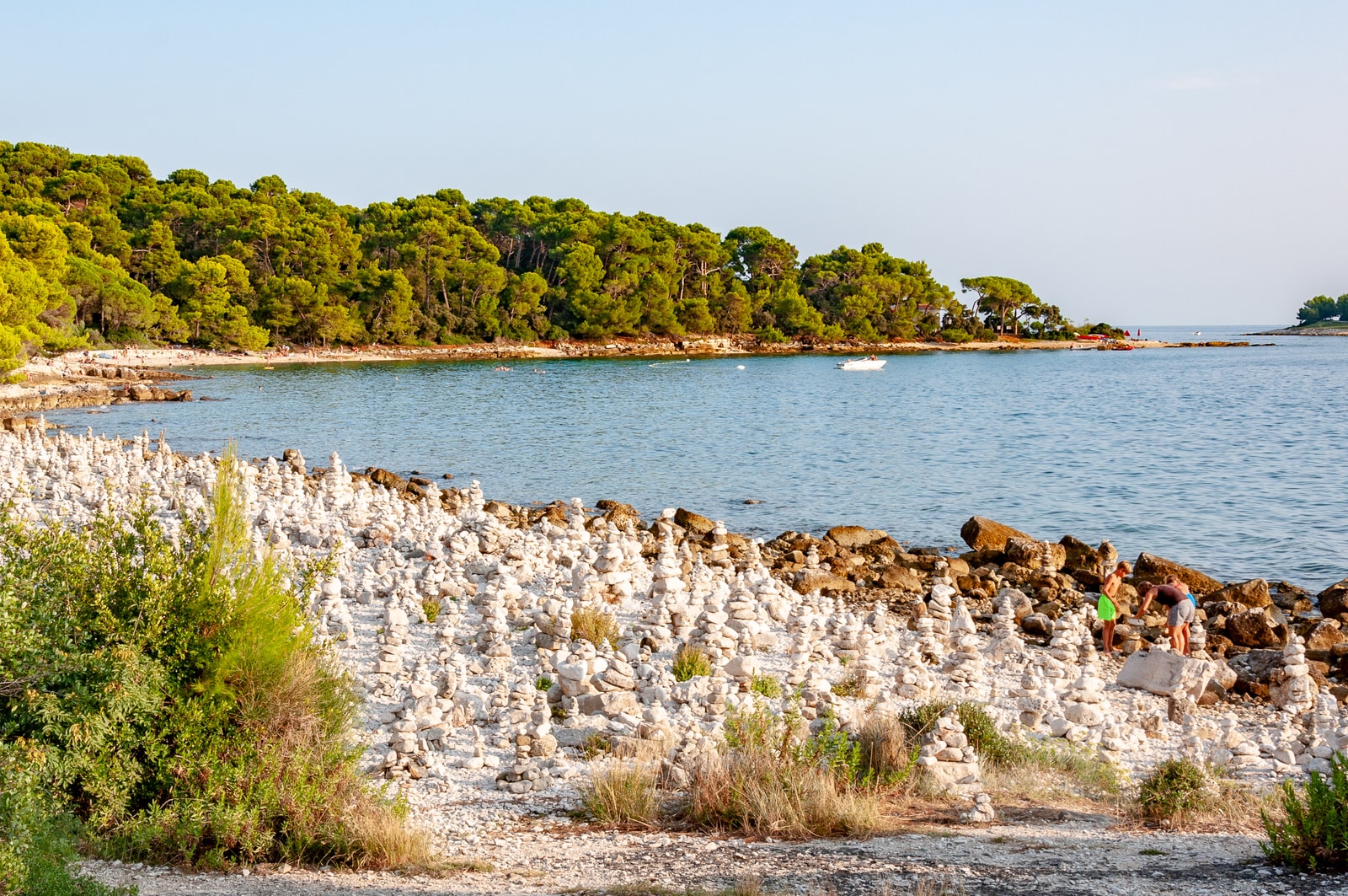 Hundreds of balanced rock stacks (cairns) cover a secluded pebble cove shore, framed by dense pine forest.