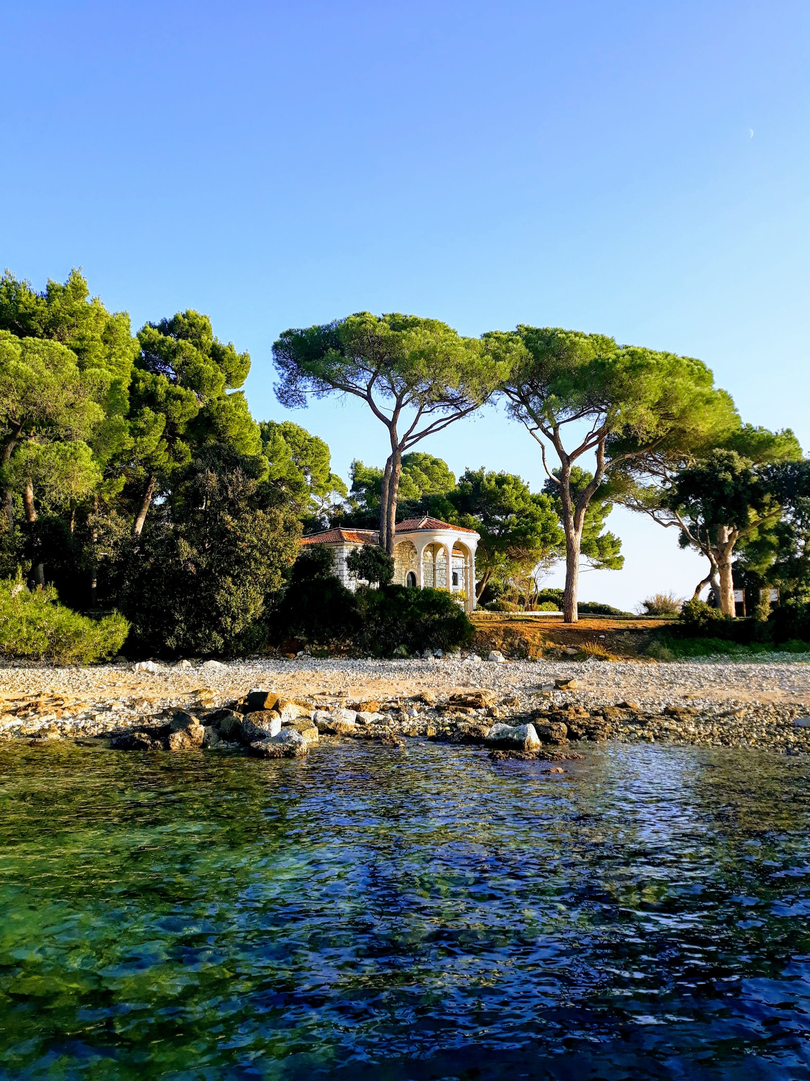 A classical-style stone pavilion with columns sits at the water's edge, surrounded by towering pine trees and crystal-clear water at Zlatni Rt.
