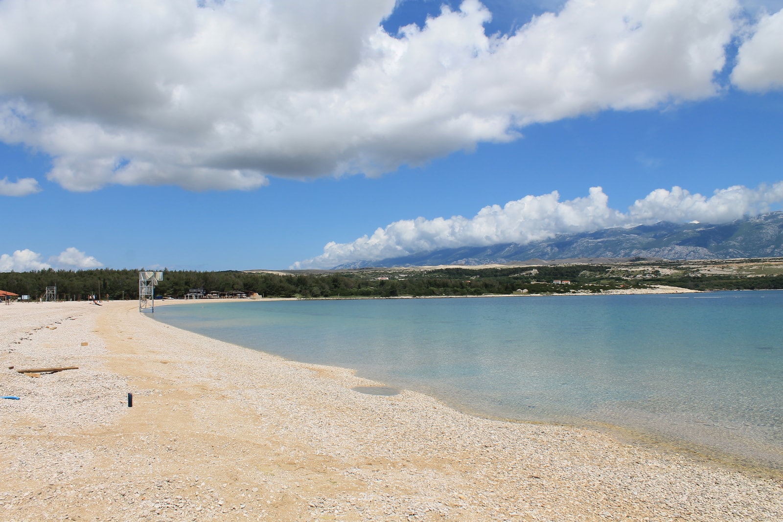 A wide view of the pebbled shoreline of Zrće Beach curving around the bay towards Kalypso Club with distant mountains.